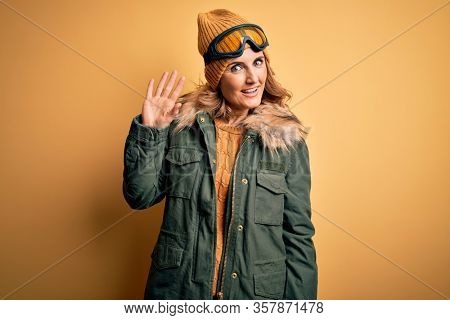 Middle age beautiful blonde skier woman wearing snow sportwear and ski goggles Waiving saying hello happy and smiling, friendly welcome gesture