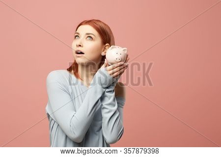 Ginger girl with piggy bank in hands standing in studio isolated on empty pink background