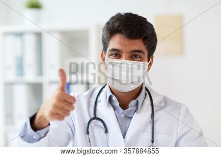 medicine, healthcare and pandemic concept - indian male doctor with stethoscope wearing protective medical mask for protection from virus showing thumbs up at clinic