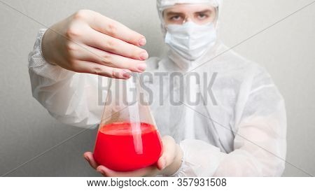 Coronovirus pandemic in the world 2020. Science against coronovirus. In the frame, a laboratory assistant in a white suit, hat and mask. Holds a flask in his hand with a red liquid coronovirus.