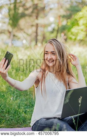 Girl Sits Outdoors And Works At A Laptop. Makes A Selfie On The Phone. Freelance. Selfeducation. The