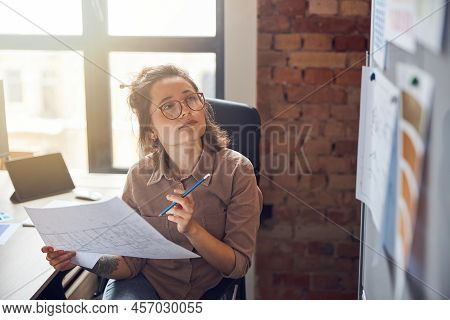 Enthusiastic Young Woman Designer Holding Blueprint Paper Looking At Board While Working On New Inte