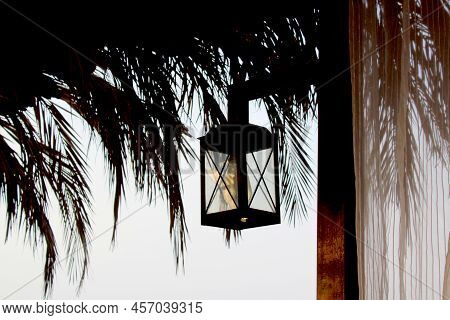 Lamp Hanging From A Wooden Pole. Jute Ceiling With Palm Leaves.evening Picture With Black Rectangula