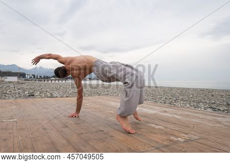Shirtless Caucasian Man Doing Backflip On Pebble Beach.