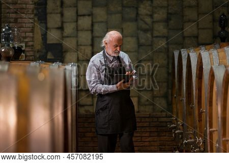 Caucasian Senior Man, A Graybeard Winemaker With Apron Checking Big Wooden Barrels In A Wine Cellar 