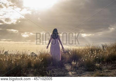 Back Of A Woman With Long Brown Hair In The Pink Linen Dress, Standing In The Open Field Facing Sky 
