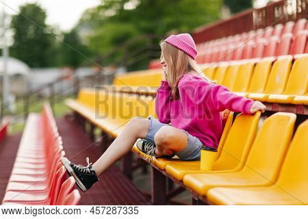 A Teenage Girl Is Sitting At The Stadium At Halftime And Talking On The Phone With Friends. Side Vie