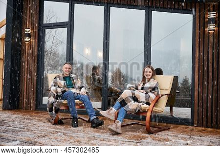 Cheerful Man And Woman Wrapped In Blanket Sitting In Chairs Outside Scandinavian House Barnhouse. Ha