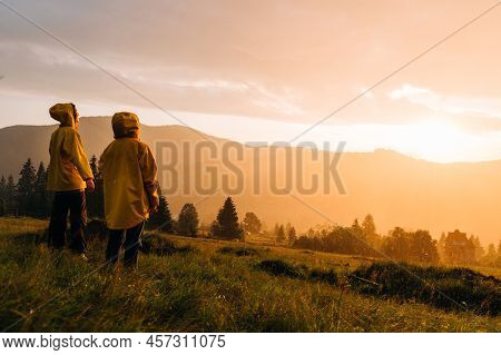 Two People In Yellow Raincoats Stand On A Meadow In The Mountains In The Rain And Watch The Unreal S