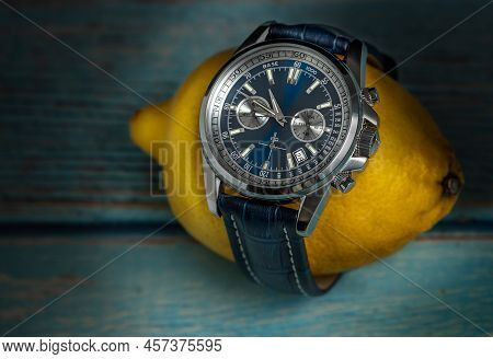 Studio Shot Of A Blue Fancy Luxury Men Wrist Watch Wrapped Around Yellow Lemon, On A Blue Wooden Tab