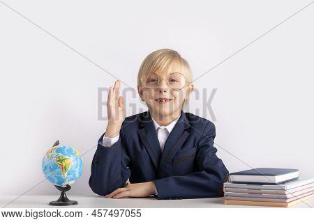 Schoolboy In Large Glasses Sits At Table With Textbooks With His Hand Raised. Boy Knows The Answer. 