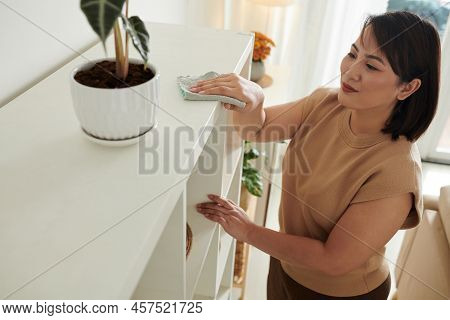 Asian Housewife Wiping Dust With Rag On The Top Of Bookcase During Housework At Home