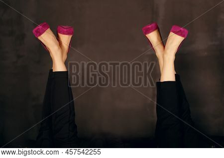 Two Pairs Of Female Legs In Lilac Shoes Upside Down On A Dark Background. Women's Legs In Bright Sho