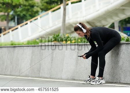 Young Sportive Girl In Wireless Headhones Tracking Her Workouts On Smartphone While Sitting Outdoors