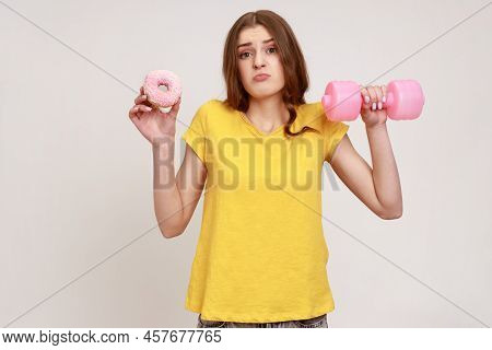 Confused Teenager Girl In Yellow Casual Style T-shirt Holding In Hands Pink Dumbbell And Donut With 