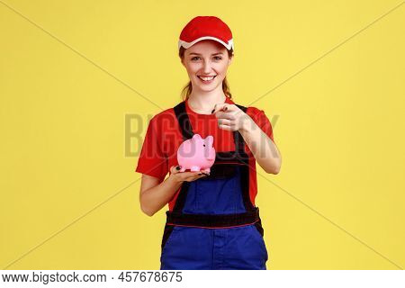 Attractive Smiling Handy Woman Standing With Piggybank And Pointing Finger To Camera, Expressing Pos