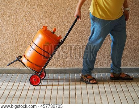 A Man On A Handcart Delivers A Gas Cylinder. Orange Propane Tank. The Head Of The Family Stocks Up F