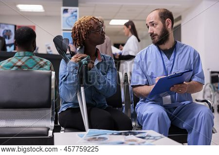 Medical Asisstant Consulting Woman With Crutches In Hospital Reception Lobby, Talking About Diagnosi