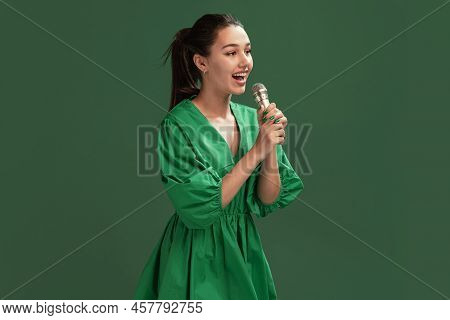 Portrait Of Beautiful Young Girl Posing In Dress, Singing In Microphone Isolated Over Green Studio B