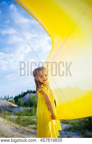 A Small 6-7 Years Old Ukrainian Girl In A Yellow Dress With Wings In Yellow Fabric By The Lake. Blue
