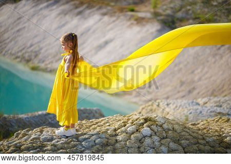A Small 6-7 Years Old Ukrainian Girl In A Yellow Dress With Wings In Yellow Fabric By The Lake. Blue