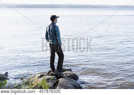 Young Man In A Cap Looking At The View Of The Lake Or River Standing On The Stones Enjoying Nature L