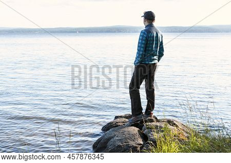 Young Man In A Cap Looking At The View Of The Lake Or River Standing On The Stones Enjoying Nature L