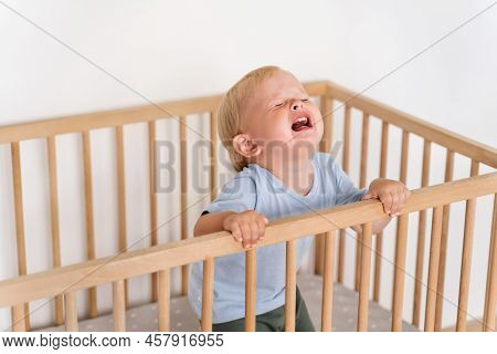 Portrait Of Upset Crying Baby Standing In Crib Getting Hysterical Seeking Attention Of Parents