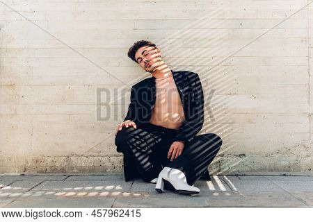 Full Body Of Feminine Male With Rhinestones On Face In High Heels Sitting Near Shabby Wall With Clos