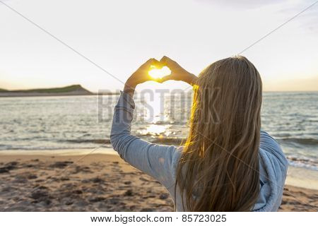 Blonde young girl holding hands in heart shape framing setting sun at sunset on ocean beach