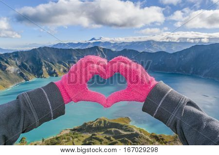 Hand with gloves forming a heart against spectacular biew of Quilotoa lake Latacunga Ecuador