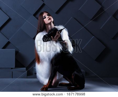 neautiful fashion woman with long hair sitting with black doberman dog on black background. She is wearing white white fur jacket