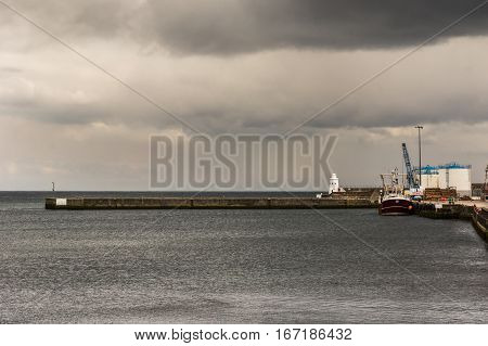 Wick Scotland - June 4 2012: The brown stone pier protrudes into Wick bay and protects the harbor of Wick. Fishing vessel and white fuel storage tanks on gray dark North Sea water under dark storm skies.