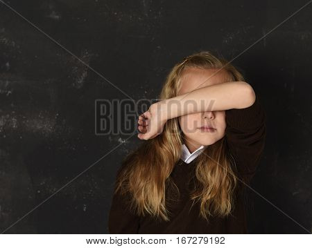 sweet young little schoolgirl in uniform covering her face with her arm crying sad victim of bullying at school isolated standing in front of dark blackboard in childhood and education problem concept