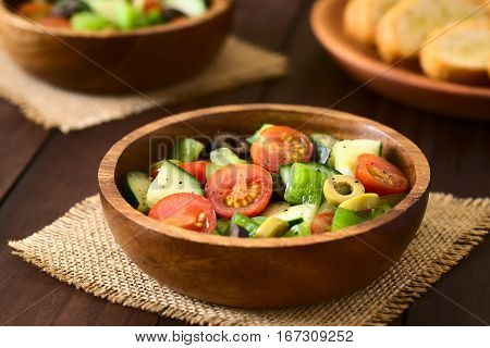 Fresh salad of black and green olives cherry tomatoes green bell pepper and cucumber seasoned with salt pepper dried oregano and basil served in wooden bowls photographed on dark wood with natural light (Selective Focus Focus in the middle of the salad)