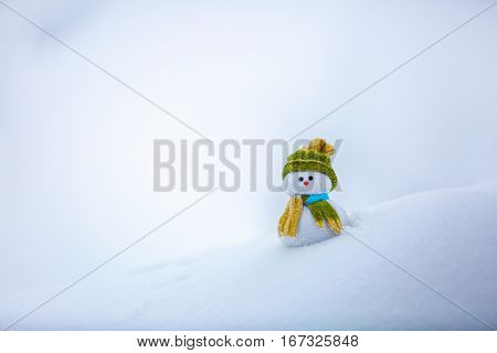 Nice dreaming snowman in green hat is standing on interesting textured snow near to the hill on cold winter day.