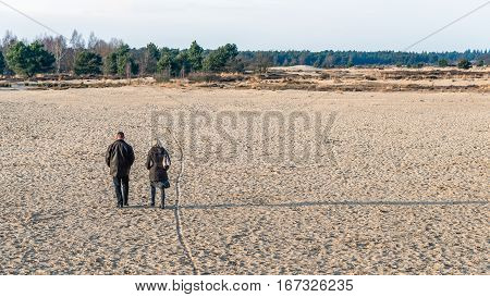 National Park in the Netherlands at the end of a clear day in the winter season. In the foreground a unknown man and woman walking in the sand. The low sun creates long shadows of them.