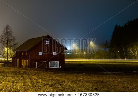 A long exposure of a old farm building at night with farmland in foreground and road with streaks of car lights in the background.