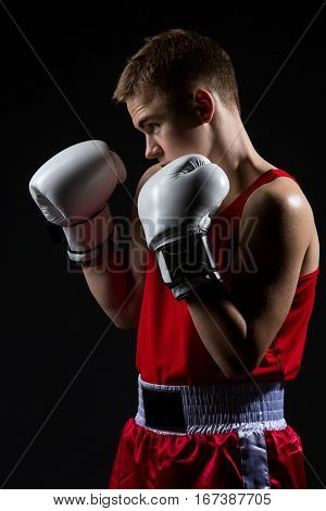 Young handsome boxer sportsman in red boxer suit and white gloves standing on black backgound. Copy space.