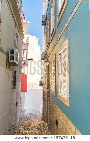 Stairs made of portuguese sidewalk rocks leading a way down to the entrance of a beautiful blue house with white metal shutters. on the other side a older facade of another house. Blue sky in the background.