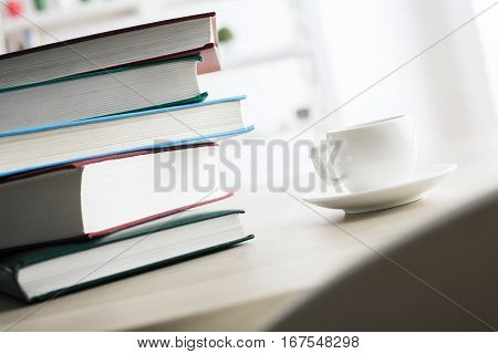 Close up of coffee cup and colorful hardcover book pile on light wooden desktop. Office workplace concept