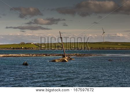 Orkneys Scotland - June 5 2012: The mast and other rusty parts of WW II warship wreck peep out of the sea near Lamb Holm Island. Wind turbine and farms on horizon. Small green band of land splits image in two. Cloudy skies. Blue sea water.