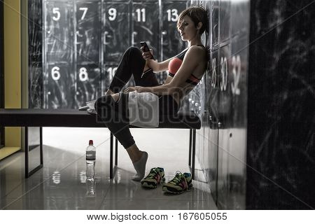 Attractive smiling girl with a mobile phone sits sideways on the bench in the locker-room in the gym. She wears black pants with white socks and a black-pink top. Horizontal.