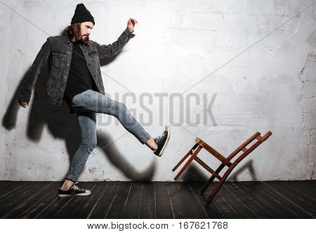 Full legth portrait of a bearded hipster man kicking chair with foot