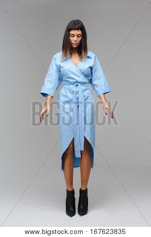 Young beautifeul woman looking down in studio with grey background. She wears a blue cloak and black boots. She behaves herself like she is standing on the brink