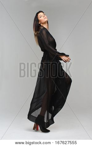 Nice model in diaphanous nightie playfully looking to the photograther camera making a step in studio with grey background. Her nightie is black, her figure is perfect