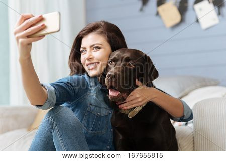 Pose like a model. Enthusiastic cheerful pretty lady taking a picture of herself hugging her nice dog while spending time in a bright blue living room