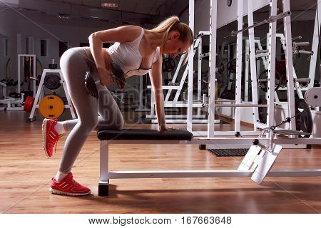 Blonde athletic woman pumping up muscles with dumbbells in gym. Side view.