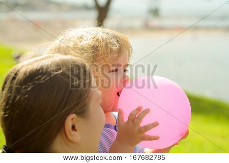 Mother and son cute baby boy with blond hair play with pink toy balloon on green grass by sea on sunny summer day on natural background at mothers day