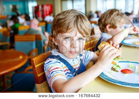 Cute healthy preschool kid boy eats hamburger sitting in school or nursery cafe. Happy child eating healthy organic and vegan food in restaurant. Childhood, health concept